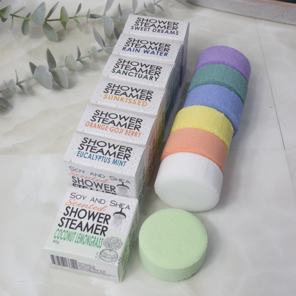 Seven boxed shower steamers labeled Soy and Shea Scented Shower Steamer are arranged next to a stack of round pastel steamer pads in purple, teal, blue, yellow, peach, and white, with one pale green steamer in front. The boxes show scents including Sweet Dreams, Rain Water, Sanctuary, Sunkissed, Orange Goji Berry, and Eucalyptus Mint.