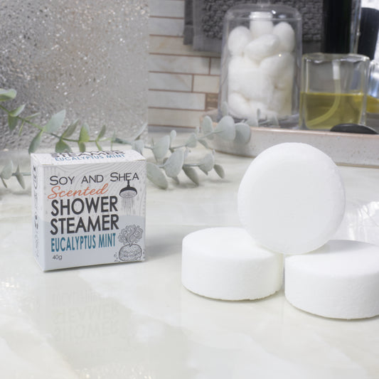Three round white shower steamer discs displayed next to their packaging box labeled 'Soy and Shea Scented Shower Steamer' in 'Eucalyptus Mint' scent, set on a white countertop with a blurred bathroom background.