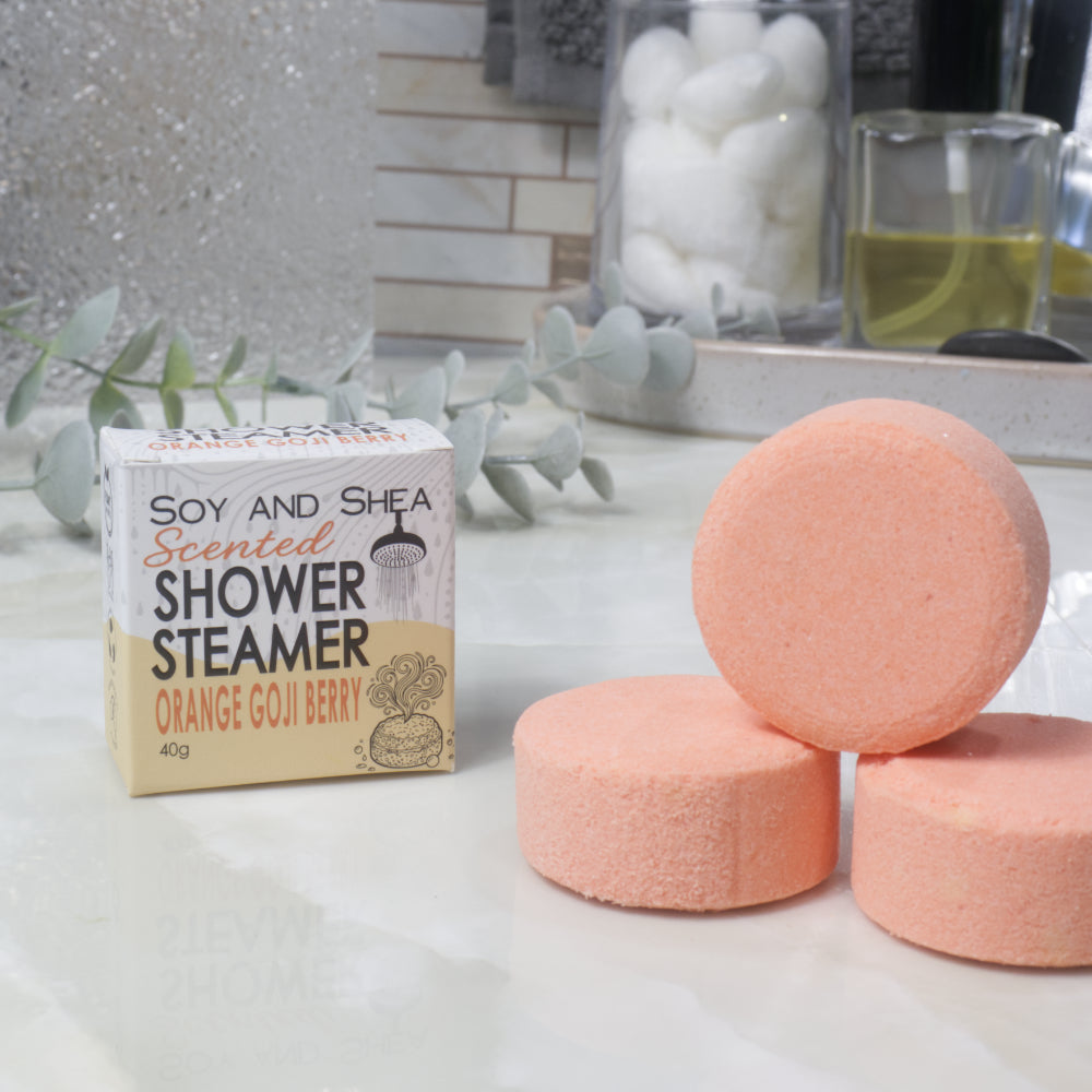 Three round orange-colored shower steamers next to their cardboard packaging box labeled 'Soy and Shea Scented Shower Steamer Orange Goji Berry' on a white countertop.