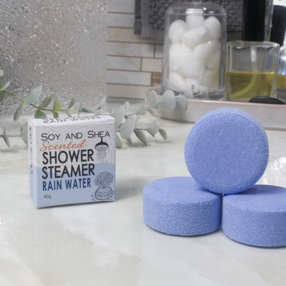 A box of Soy and Shea Scented Shower Steamer Rain Water sits next to three round blue shower steamers on a light marble countertop with spa-style props in the background.