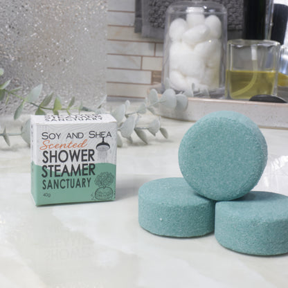 Three round dark green shower steamer discs displayed on a white marble surface next to their branded cardboard box, with decorative eucalyptus leaves and bathroom accessories blurred in the background.