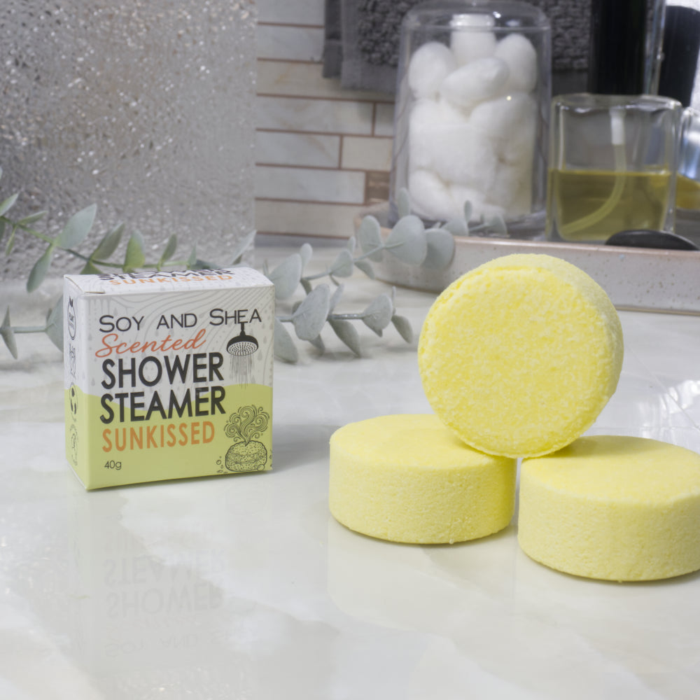A box of Soy and Shea Scented Shower Steamer in 'Sunkissed' alongside three round, yellow shower steamers on a white marble countertop with decorative eucalyptus leaves and bathroom accessories in the background.