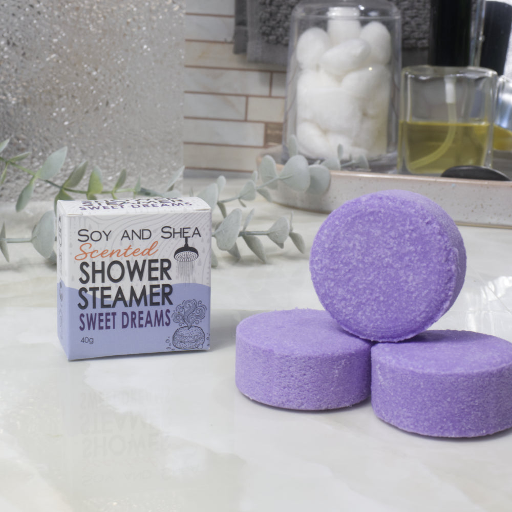 Three purple shower steamer discs are arranged on a white marble-like surface next to their product box labeled 'Soy and Shea Scented Shower Steamer Sweet Dreams'. The background shows a softly blurred bathroom setting with decorative leaves and jars.