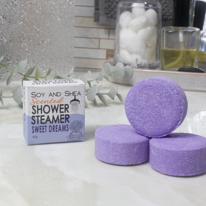 Three purple shower steamer discs are arranged on a white marble-like surface next to their product box labeled 'Soy and Shea Scented Shower Steamer Sweet Dreams'. The background shows a softly blurred bathroom setting with decorative leaves and jars.