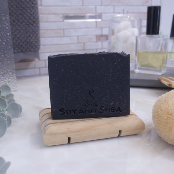 A black bar of soap with the text 'SOY & SHEA' stamped on it, resting on a wooden soap dish on a bathroom countertop.