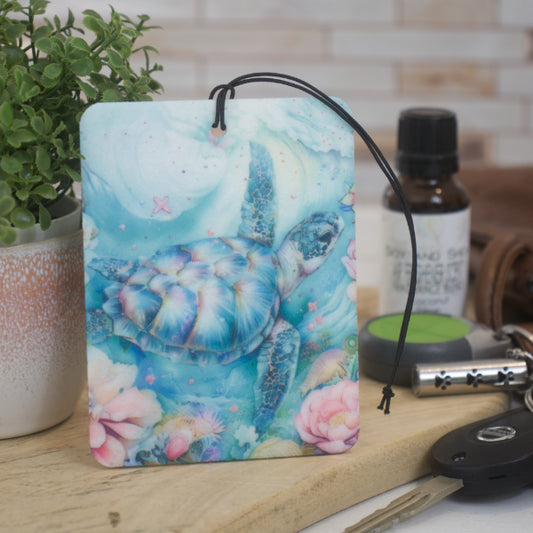 A car air freshener featuring an artistic depiction of a turtle underwater surrounded by floral elements, placed on a wooden surface with various items in the background.  