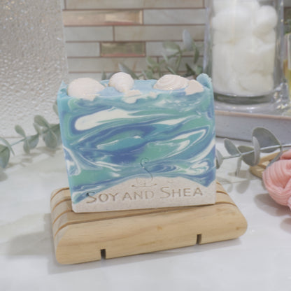 A soap bar sits on a wooden soap rack on a bathroom counter next to a jar of cotton balls.   The soap has a beige base layer topped with a marbled blue and white top layer made to look like water.   The top is decorated with beige seashells