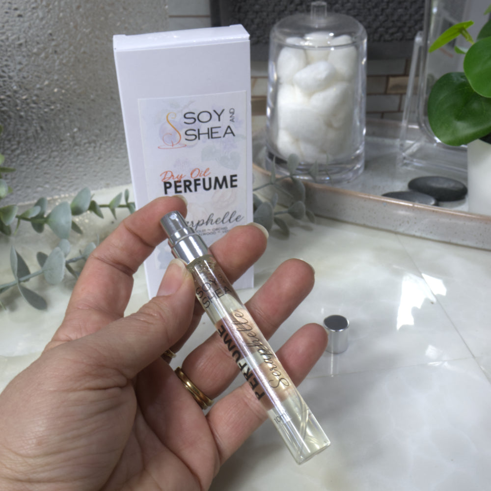 A hand holds a small clear glass perfume sample vial with a silver atomizer cap in front of its white retail box labeled 'Soy Shea' and 'Dry Oil Perfume'. The scene is set on a light marble-like surface with a glass jar of cotton pads and greenery in the background.