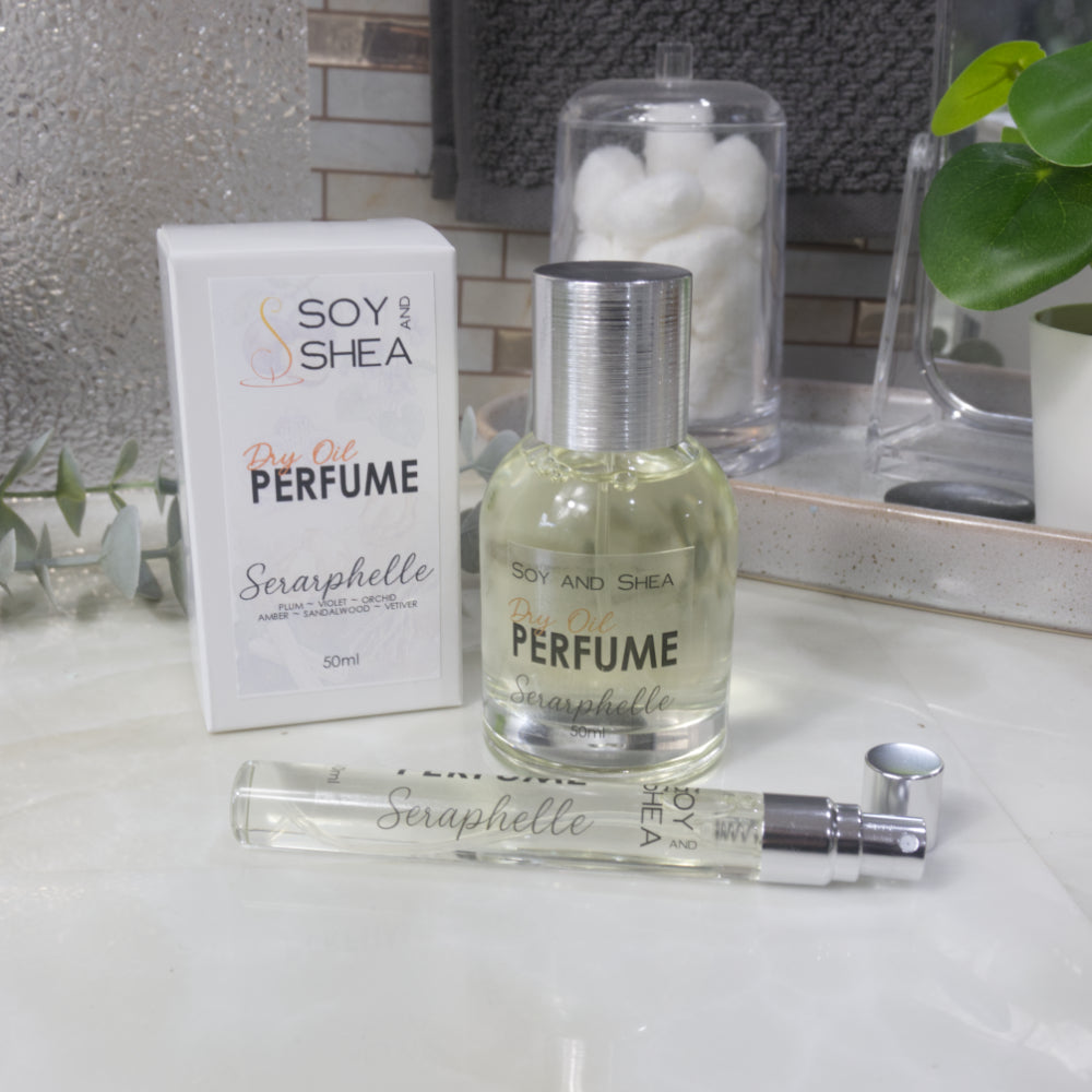 A selection of perfume bottles are presented on a bathroom countertop.  To the front is a clear, narrow, tall bottle labeled as 10ml with a silver cap and behind is a clear round, short bottle with silver lid labelled as 50ml.  Behind sits a white box with label identifying as Dry Oil Perfume Seraphelle.
