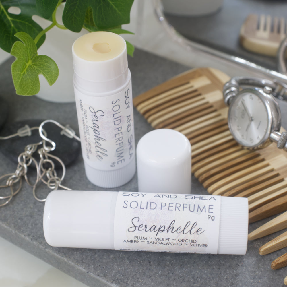 Two white tubes of Solid Perfume are displayed on a grey stone tray along with silver earrings, watch and greenery.   One tube lays flat  displaying the fragrance notes while another tube stands upright behind it with the lid removed and to the right revealing a beige balm