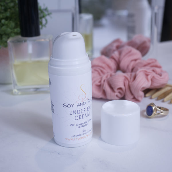 A white bottle of Under Eye Cream sits on a bathroom counter along with a scrunchie, small mirror, bottle of perfume, a ring and plant.  The white lid has been removed and sits on the counter revealing a serum pump top.