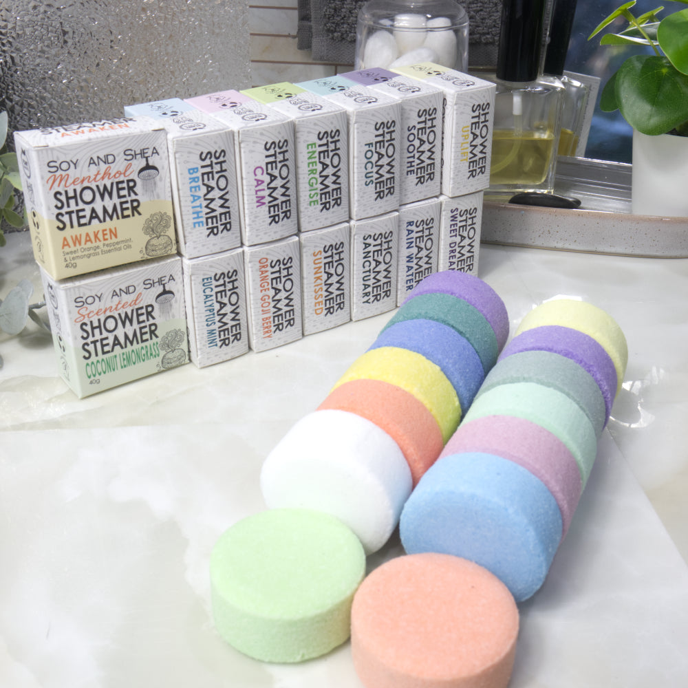 A set of colorful round shower steamer discs in the foreground with their matching branded boxes lined up in the background on a bathroom counter.