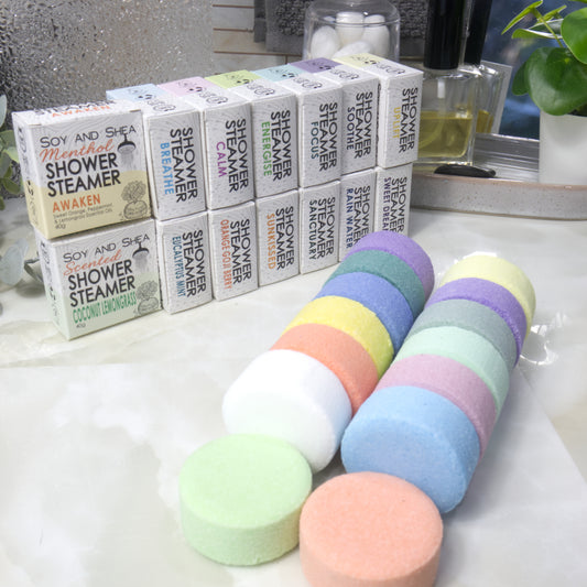 A set of colorful round shower steamer discs in the foreground with their matching branded boxes lined up in the background on a bathroom counter.