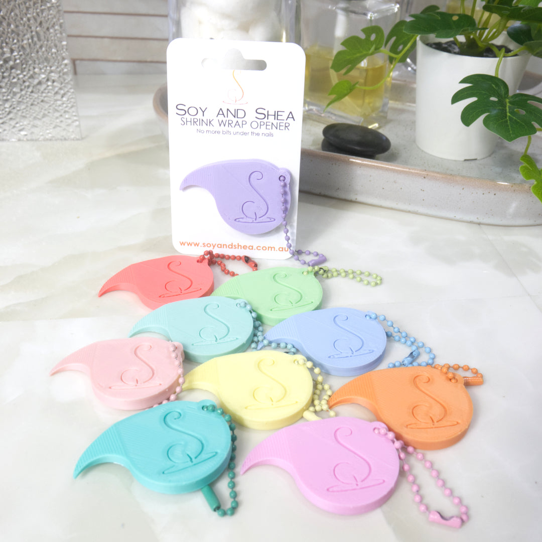 A collection of 3D printed Shrinkwrap openers lay on a marble bathroom counter in an arrange of pastel colours.  Each opener has a matching ball and chain keyring on it.  A lilac opener is attached to a white presentation card and stands upright behind the other openers.  In the background is a plant and a bottle of perfume