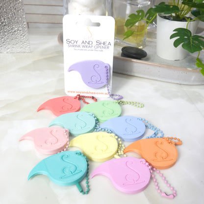 A collection of 3D printed Shrinkwrap openers lay on a marble bathroom counter in an arrange of pastel colours.  Each opener has a matching ball and chain keyring on it.  A lilac opener is attached to a white presentation card and stands upright behind the other openers.  In the background is a plant and a bottle of perfume