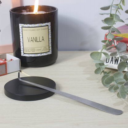 A black glass jar candle with a lit flame and a vanilla label sits on a light wood table beside a black metal lid and long metal tool, with books and eucalyptus leaves in the background.  the metal tool is long, narrow and thin and features a hooked end.