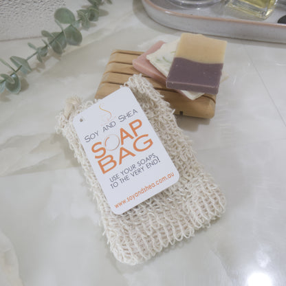  A rough cotton like bag with a swing tag reading SOAP BAG lies on a marble surface with the top resting on a wooden soap dish with a few small pieces of soap.
