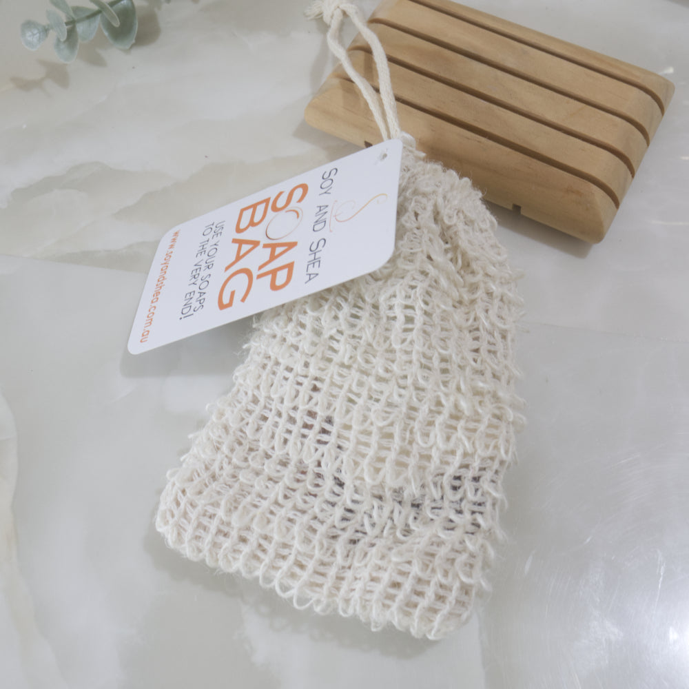 A cream loosely woven cotton bag with a swing tag identifying it as a SOAP BAG lies on a marble surflace next to wooden soap dish