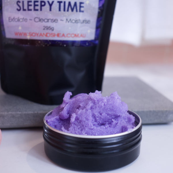 A zoomed in image of a black tin filled with purple sugar scrub sitting on a white bench with the blurred image of the packaging in the background.
