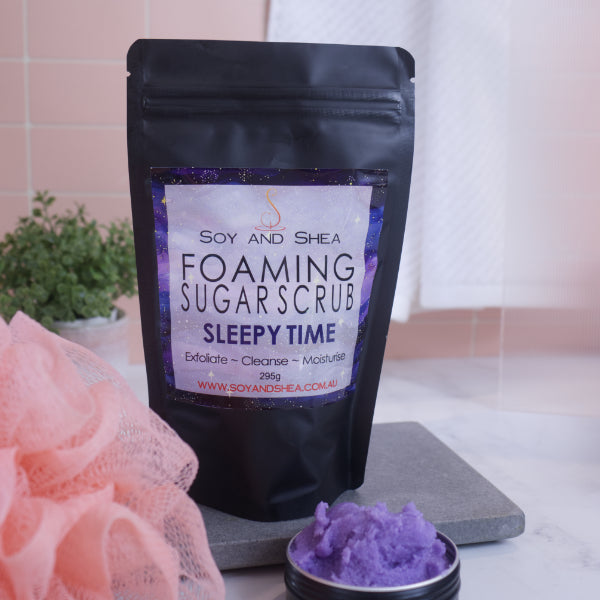 A black zip lock pouch stands up-right on a grey stone tray in front of pink tiled wall.  The Label reads  FOAMING SUGAR SCRUB SLEEPY TIME against a dark galaxy background.    In front is a small black tin overflowing with a purple scrub .
