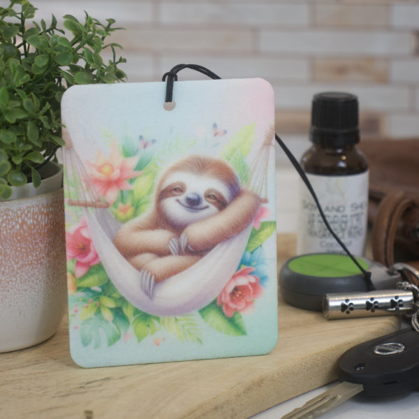 A car air freshener rests against a potted plant and next to a set of keys.  The air freshener depicts a sloth in a hammock surrounded by tropical flowers.