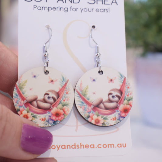 A hand holds a white card branded with SOY AND SHEA with a pair of earrings hanging on hooks.  The earrings are round with an illustration of a sloth in a red hammock over tropical flowers with butterflies fluttering above