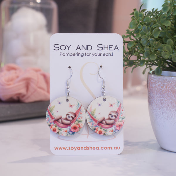A pair of earrings on silver hooks hang on a white card branded with SOY AND SHEA leaning against a pot with a green leafy plant and a blurred background.  The earrings are round with an illustration of a sloth lying in a red hammock above a bed of tropical flowers with butterflies above.