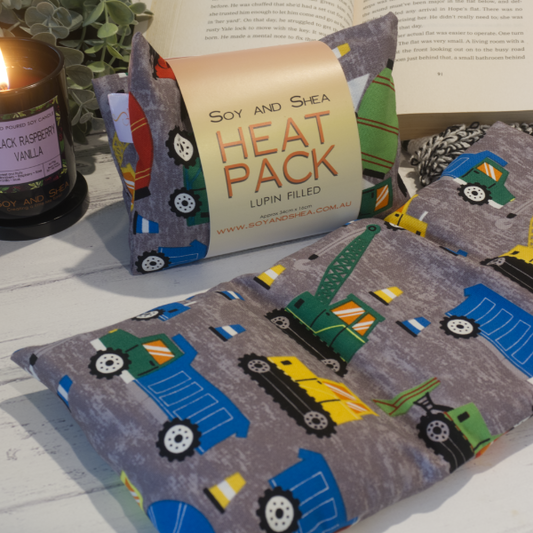 Two Small Heat packs are displayed on a wooden bench, with one laying out flat while the other sits behind with a belly band label reading HEAT PACK LUPIN FIlled.   An open book, burning candle and greenery can be seen in the background evoking a cosy feel.  The heat pack is a dark grey mottled colour with various construction vehicles in blue, green and yellow along with some traffic cones.