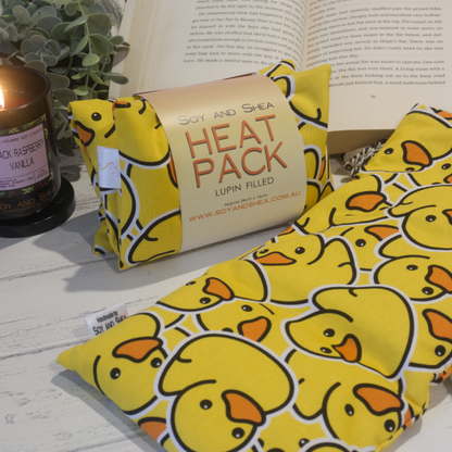 Two Small Heat packs are displayed on a wooden bench, with one laying out flat while the other sits behind with a belly band label reading HEAT PACK LUPIN FIlled.   An open book, burning candle and greenery can be seen in the background evoking a cosy feel.    The Heat packs are a bright yellow featuring yellow rubber ducks stacked upon each other.