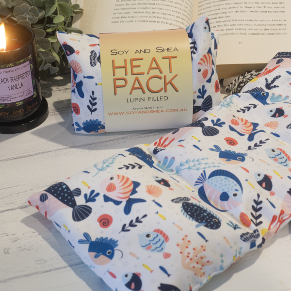 Two Small Heat packs are displayed on a wooden bench, with one laying out flat while the other sits behind with a belly band label reading HEAT PACK LUPIN FIlled.   An open book, burning candle and greenery can be seen in the background evoking a cosy feel.    The Heat packs features a variety of illustrated reef fish in shades of blue with orange highlights swimming between coral pieces on a white background