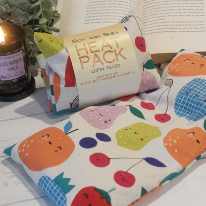 Two Small Heat packs are displayed on a wooden bench, with one laying out flat while the other sits behind with a belly band label reading HEAT PACK LUPIN FIlled.   An open book, burning candle and greenery can be seen in the background evoking a cosy feel.    The Heat packs have a cream fabric covered with stylised fruits including lemon, pear, orange, cherry and blueberries.  All the fruits hava  smiling kawai face