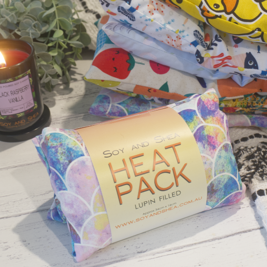 A small heat pack with pastel pink and blue mermaid scales and wrapped with a label saying HEAT PACK LUPIN FILLED sits on a wooden top with a stack of other small heat packs behind it showing various fabric designs. A glowing candle and plant sit in the background.