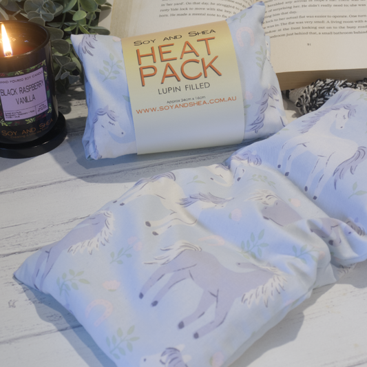 Two Small Heat packs are displayed on a wooden bench, with one laying out flat while the other sits behind with a belly band label reading HEAT PACK LUPIN FIlled.   An open book, burning candle and greenery can be seen in the background evoking a cosy feel.    The Heat packs are a pale blue colour with illustrations of horses in light purple and white prancing between green leaves and pink horseshoes