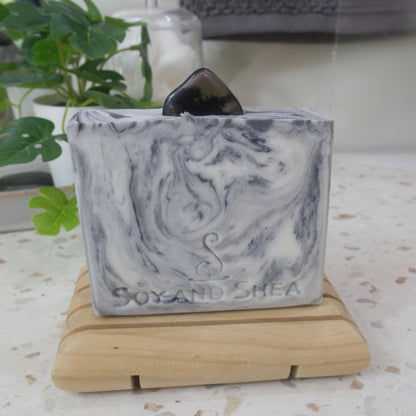 A soap bar stands on a wooden soap rack on a bathroom counter top with a plant in the background.  The soap has a white base with intricate swirls of black to grey and features a Smoky Obsidian Stone on the top.