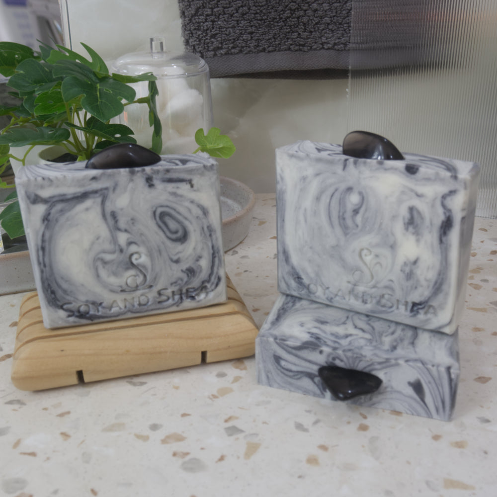 A selection of soaps are displayed on a bathroom countertop along with a oplant.  One soap bar stands on a wooden soap rack to the back left, while another lies on its side to the right front showcasing the Smoky Obsidian stone on the top.  A third soap stands on this.  All soaps have a base of white colour with intricate swirls of grey to black.  Each soap has a stone on the top.