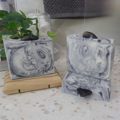 A selection of soaps are displayed on a bathroom countertop along with a oplant.  One soap bar stands on a wooden soap rack to the back left, while another lies on its side to the right front showcasing the Smoky Obsidian stone on the top.  A third soap stands on this.  All soaps have a base of white colour with intricate swirls of grey to black.  Each soap has a stone on the top.