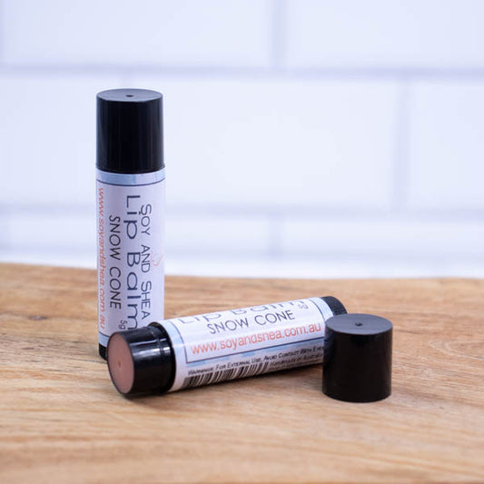 Two black tubes of lip balm are displayed on a wooden surface with blurred tiled background.  One tube stands on its end while the other lays flat in front with the cap removed revealing a light red balm.  The label has a blue background.