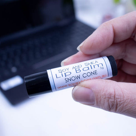 A hand holds a black tube of lip balm against a blurred background with an open laptop computer on a white surface.  The label has text LIP BALM SNOW CONE on a white background with pale blue border.