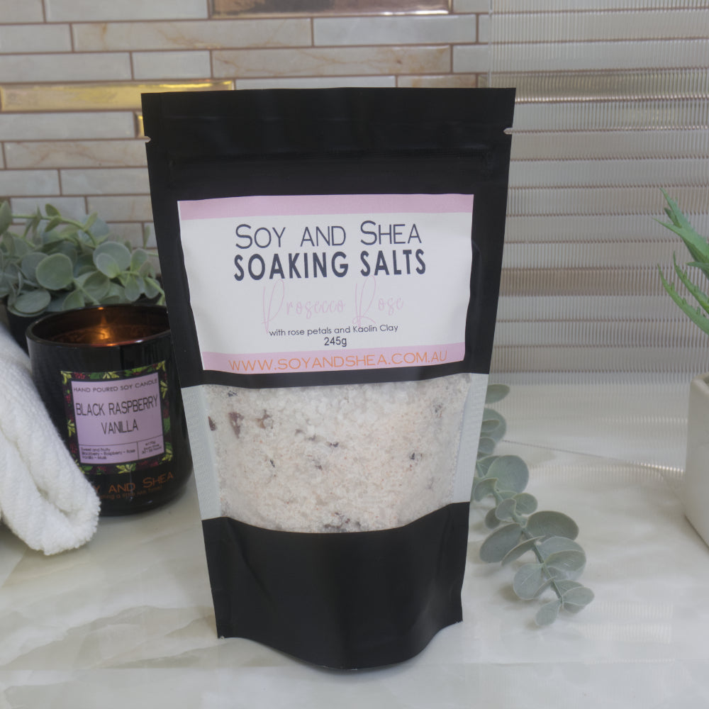 A black Soy and Shea Prosecco Rose Soaking Salts pouch sits on a bathroom counter with a candle, towel, and eucalyptus. White and pink bath salts are visible through the pouch’s clear window.