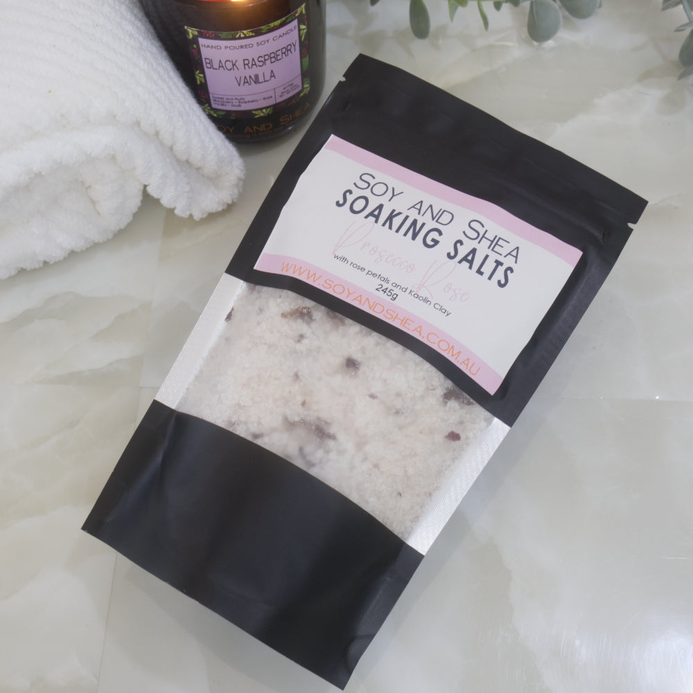 A black pouch of Soy and Shea Soaking Salts with pink label lies on a marble bench next to a towel 