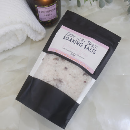 A black pouch of Soy and Shea Soaking Salts with pink label lies on a marble bench next to a towel 