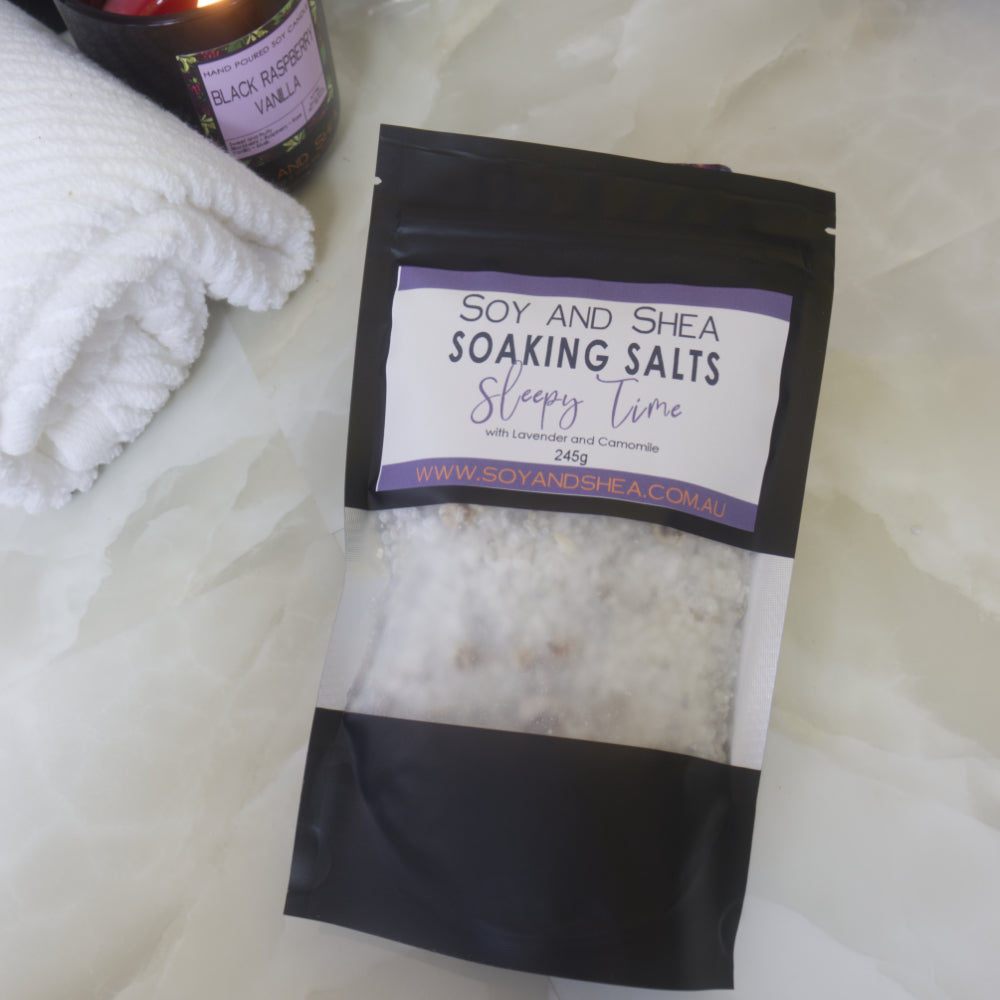 A black pouch of Soy and Shea Soaking Salts with purple label lies on a marble bench next to a towel and lit candle