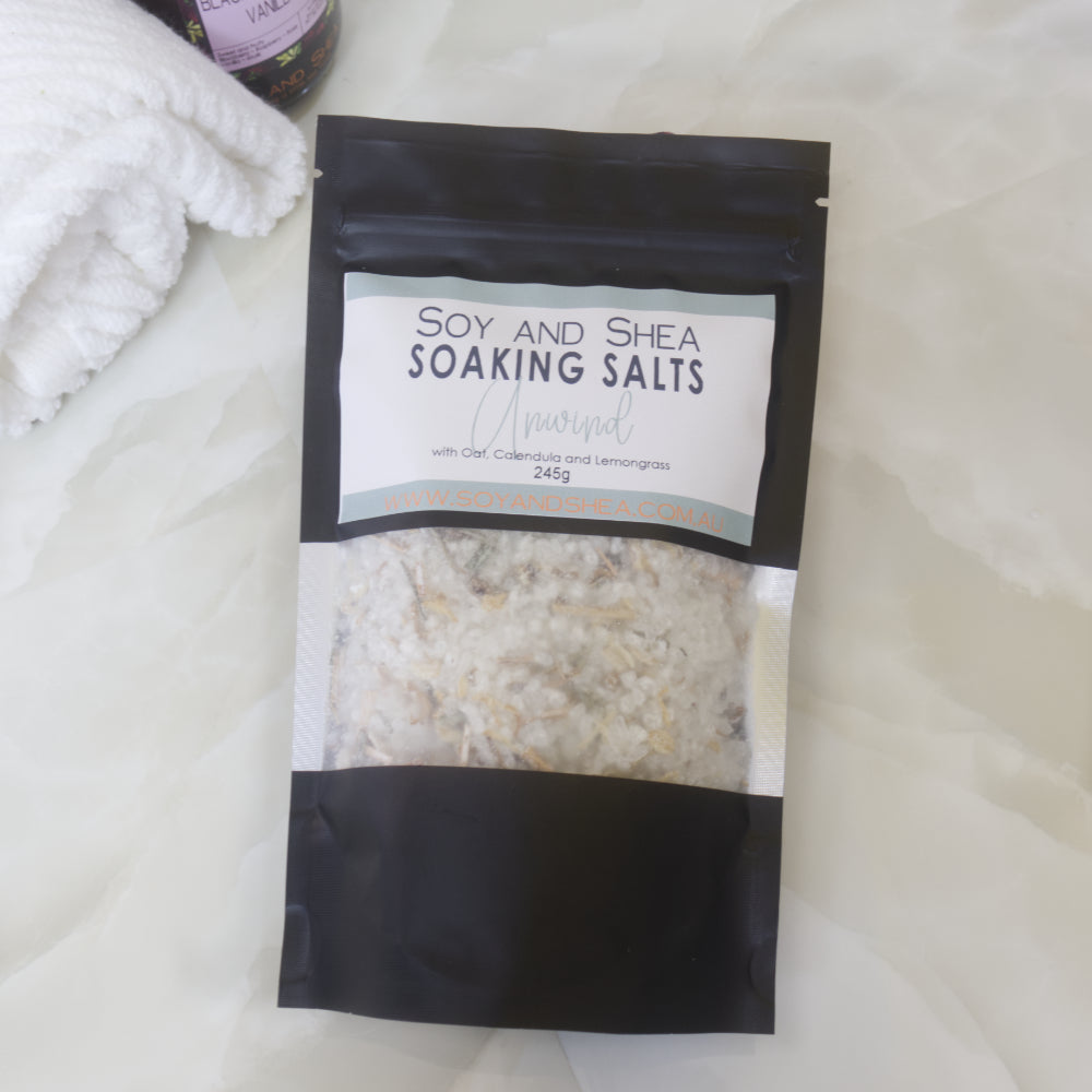 A black pouch of Soy and Shea Soaking Salts with pale green label lies on a marble bench next to a towel