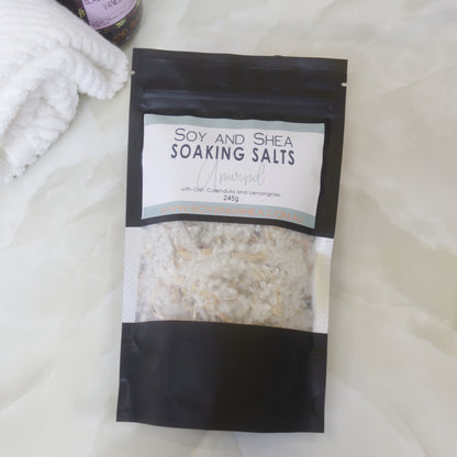 A black pouch of Soy and Shea Soaking Salts with pale green label lies on a marble bench next to a towel