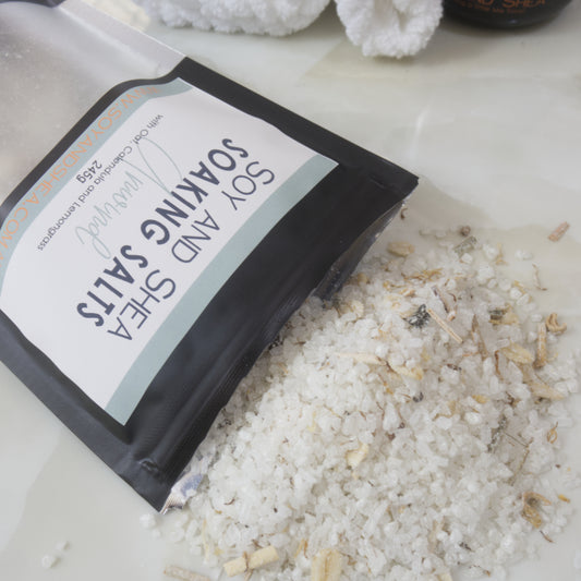 A black pouch of Soy and Shea Soaking Salts with a pale green label spilling salt across a marble bench