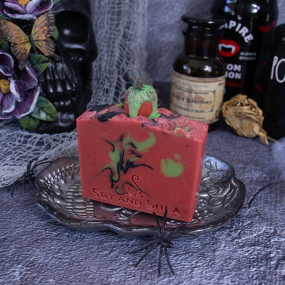 A rectangular bar of Poison Apple Soap Bar (Halloween Edition) by Soy and Shea, featuring Halloween-themed designs like a green pumpkin on top, sits on a silver plate adorned with plastic spiders. Infused with hints of cinnamon, the soap is surrounded by dark, gothic-style bottles and decorations, including a skull and cobwebs.