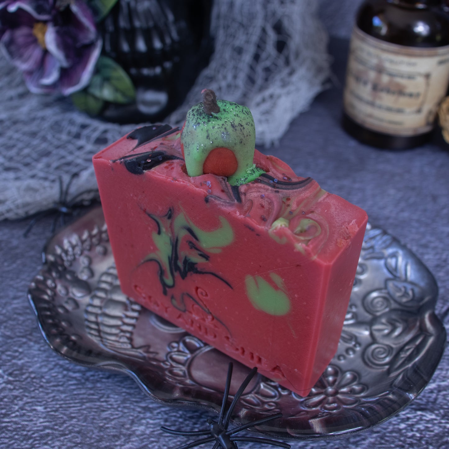 The "Poison Apple Soap Bar (Halloween Edition)" by Soy and Shea is a rectangular red handmade soap featuring black and green swirls and a hint of cinnamon fragrance, topped with a green apple decoration. It sits on a decorative silver plate amidst Halloween-themed decor, including a small black spider and potion bottles.