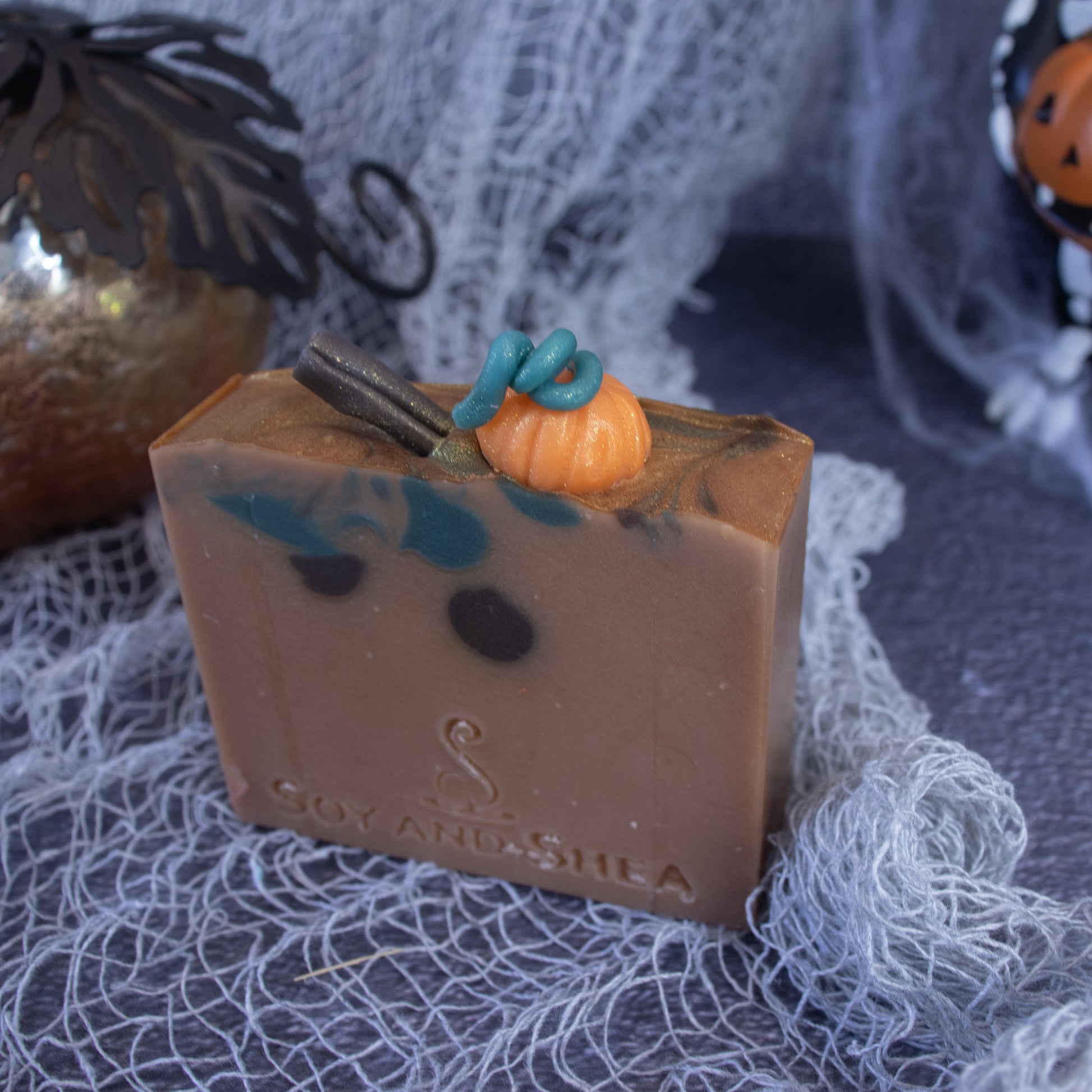 A Spiced Pumpkin Soap Bar (Halloween Edition) by Soy and Shea, featuring rustic brown hues with intricate swirls of dark and teal colors, sits elegantly atop a gray textured cloth. The soap is beautifully adorned with a small pumpkin and stick decoration on top, exuding a delightful hint of vanilla. It is embossed with "Soy and Shea," set against a backdrop of fall-themed decor.