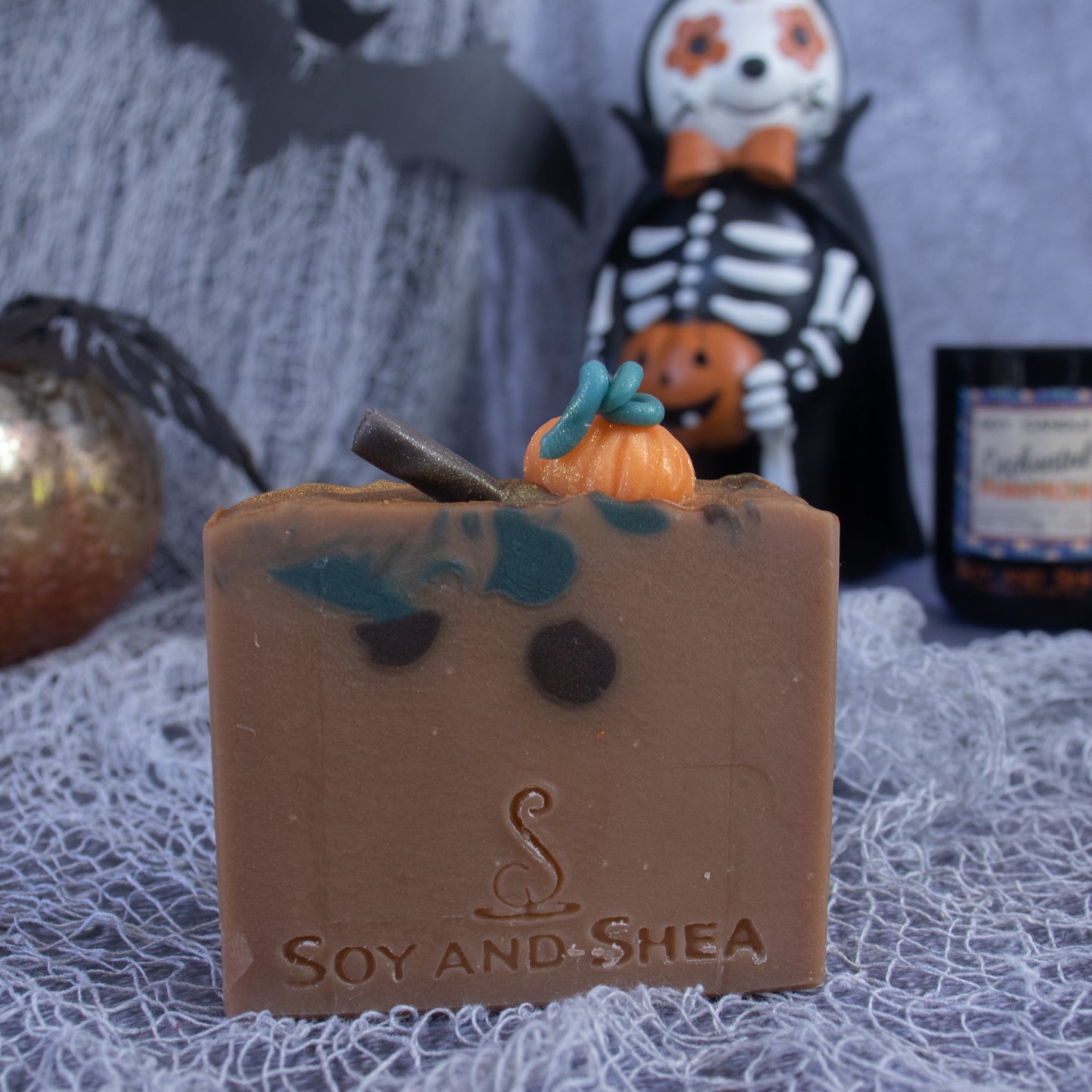 A handmade Spiced Pumpkin Soap Bar from Soy and Shea showcases a decorative pumpkin and bat design. Resting on textured gray fabric, it is surrounded by Halloween props such as a skeleton figurine and a silver pumpkin, subtly infused with the sweet scent of vanilla.