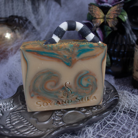 The Sandworm Soap Bar (Halloween Edition) by Soy and Shea, designed to resemble a handbag with swirled teal and gold accents, rests on an ornate soap dish. The backdrop is adorned with Halloween-themed elements such as a skull and cobwebs. This handmade soap is ideal for infusing your Halloween decor with a sense of fun and luxury.